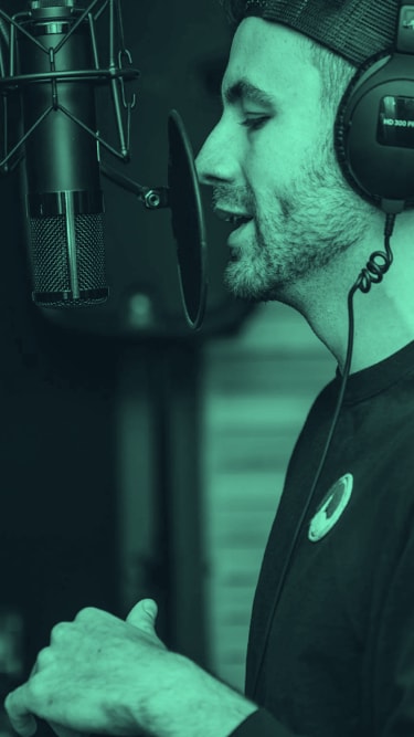 podcast host talking into a microphone while wearing earphones and a backwards hat